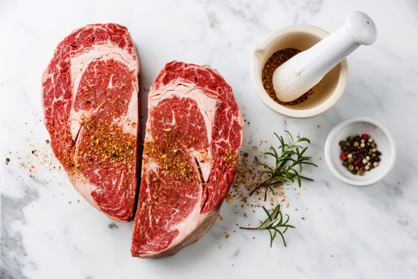 Two raw, seasoned ribeye steaks on a marble surface, next to a mortar with ground spices, a sprig of rosemary, and a bowl of mixed peppercorns.