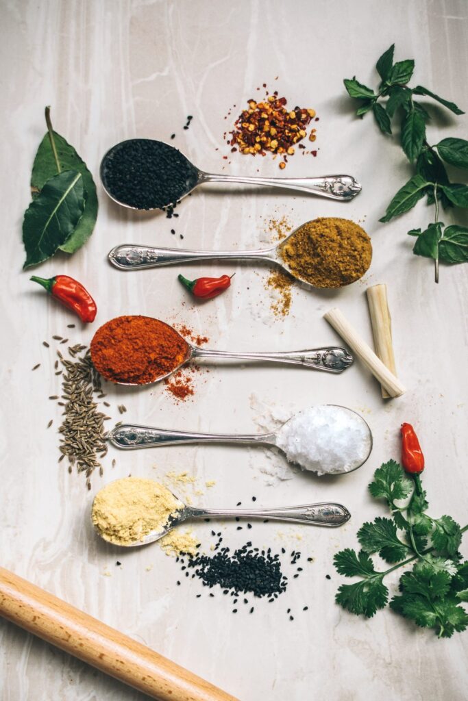 Six spoons filled with various spices and herbs are arranged in a vertical row on a light surface, accompanied by fresh herbs, chili peppers, and a wooden kitchen tool.