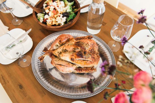 A roasted chicken on a silver platter, surrounded by a salad bowl, a bottle of water, glass of water, and plates on a wooden table. Flowers are partially visible in the foreground.