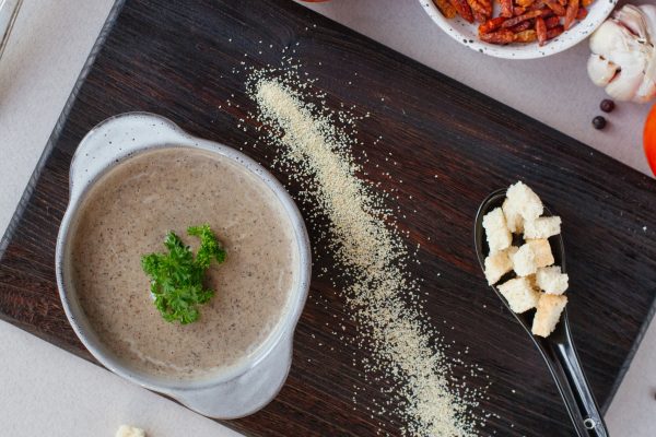 A bowl of creamy mushroom soup garnished with parsley is on a wooden board, accompanied by a spoon with croutons and a sprinkle of grated cheese.