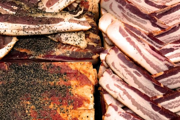 An assortment of uncooked bacon slices arranged in piles, seasoned with black pepper on the left and plain on the right.