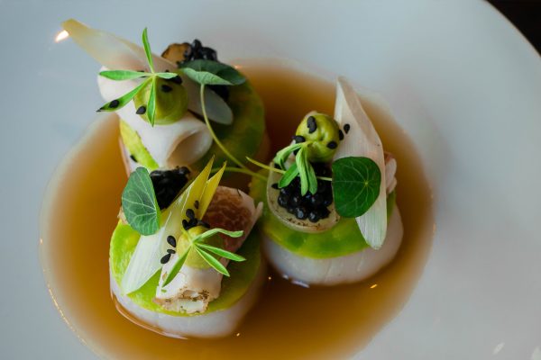 A plate with three gourmet appetizers, each featuring thinly sliced vegetables, garnishes, and black caviar, served in a light broth.